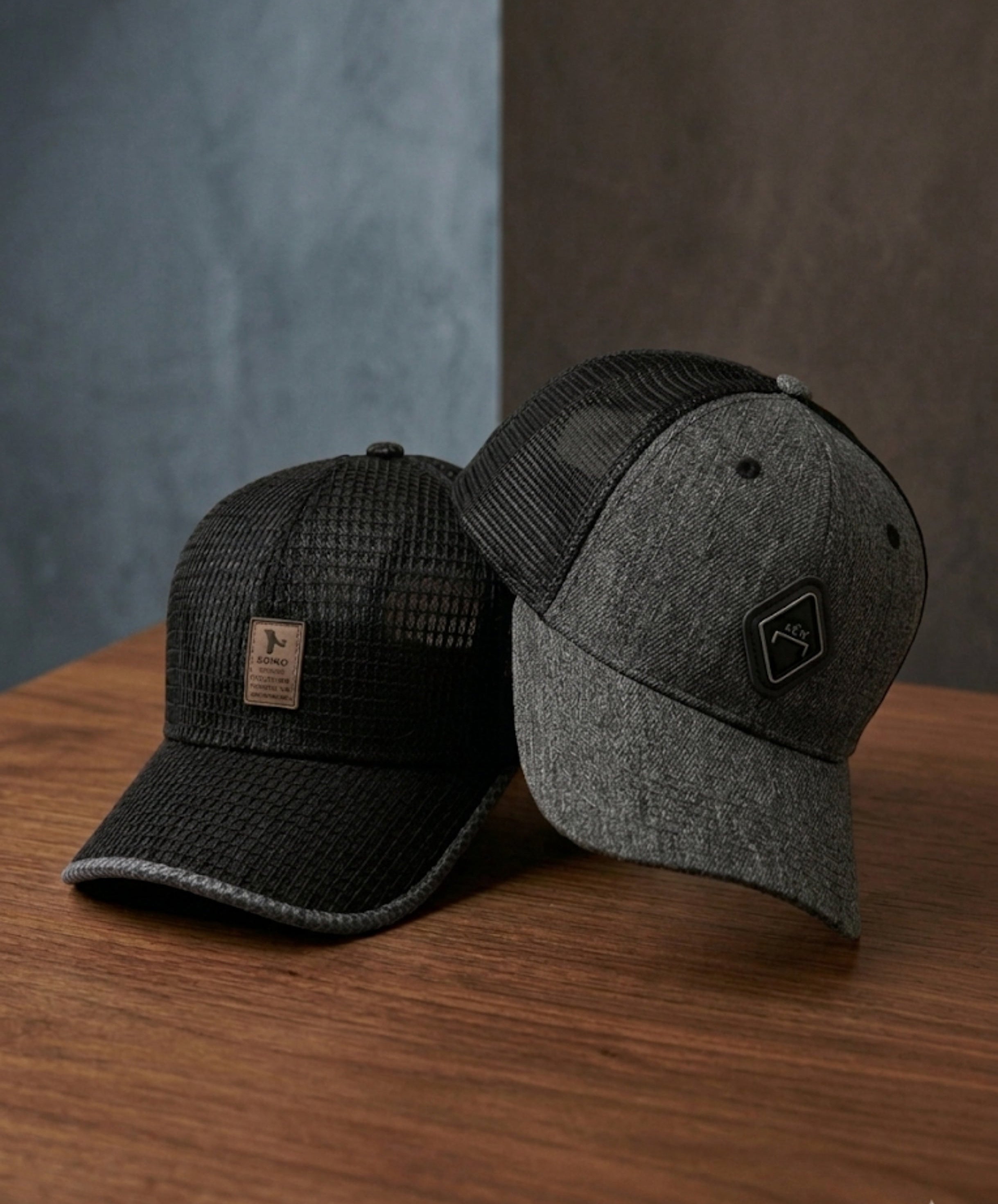 Two trucker-style baseball caps displayed on a wooden surface against a split gray and brown background. One cap is black with a textured front panel and black mesh back, featuring a small tan rectangular patch on the front.